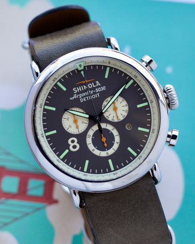 Shinola watch with a dark face and brown leather strap on a light blue background