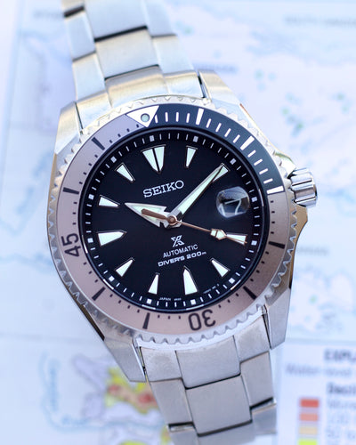 Seiko watch with black dial and silver metal strap on a blurred map background