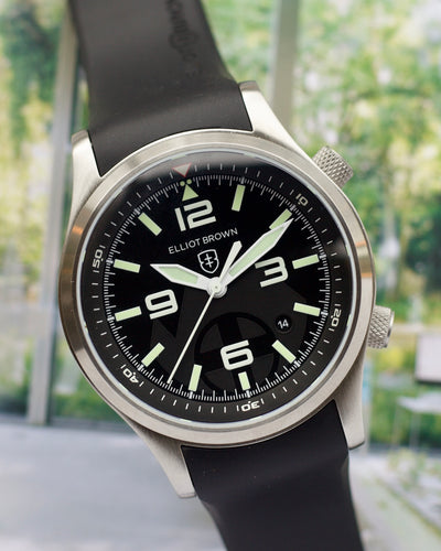 Watch with black strap and black dial featuring brand name, blurred green outdoor background